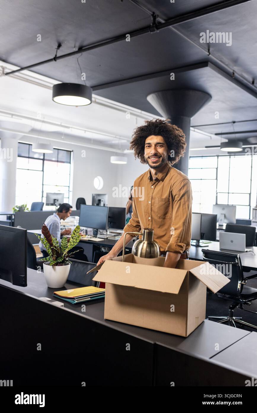 Uomo sorridente che disimballava la scatola alla moderna scrivania dell'ufficio, iniziando un nuovo lavoro Foto Stock