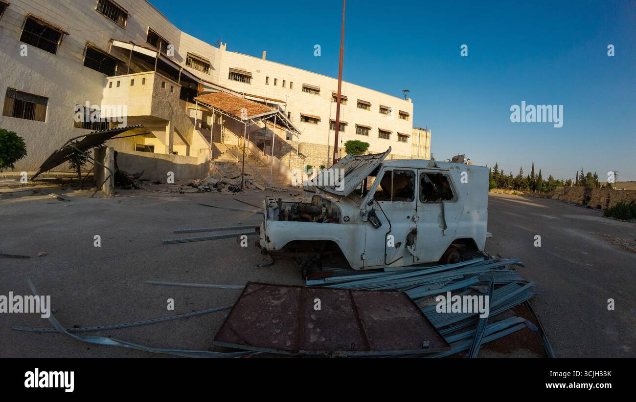 Furgone abbandonato e danneggiato di fronte a un edificio abbandonato sotto il cielo azzurro. Siria, prigione di Sednaya. Foto Stock
