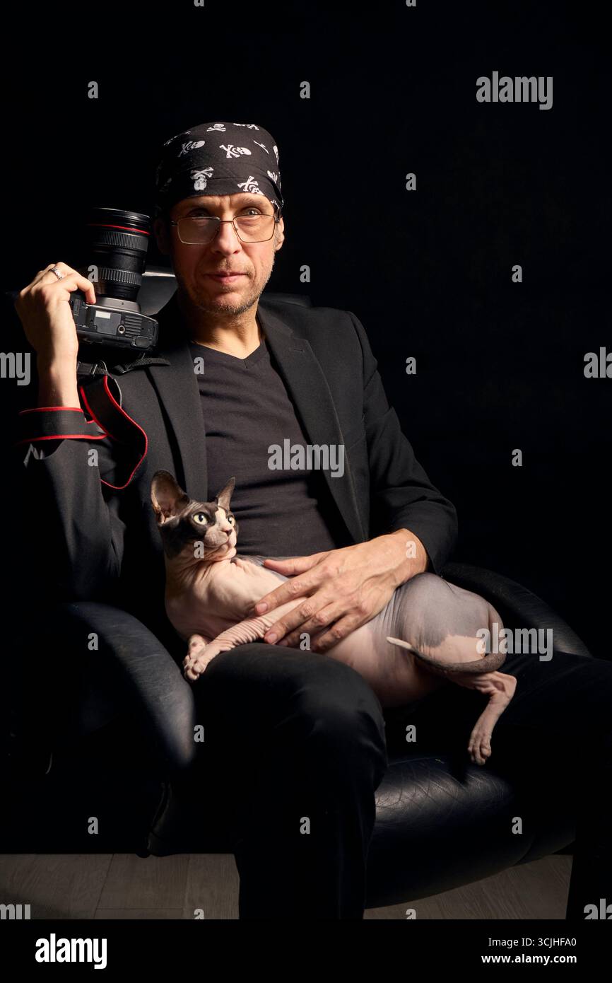 Un ritratto creativo di un fotografo in abiti neri con un gatto Sphynx in grembo, che tiene una macchina fotografica in uno studio buio. L'immagine riflette l Foto Stock