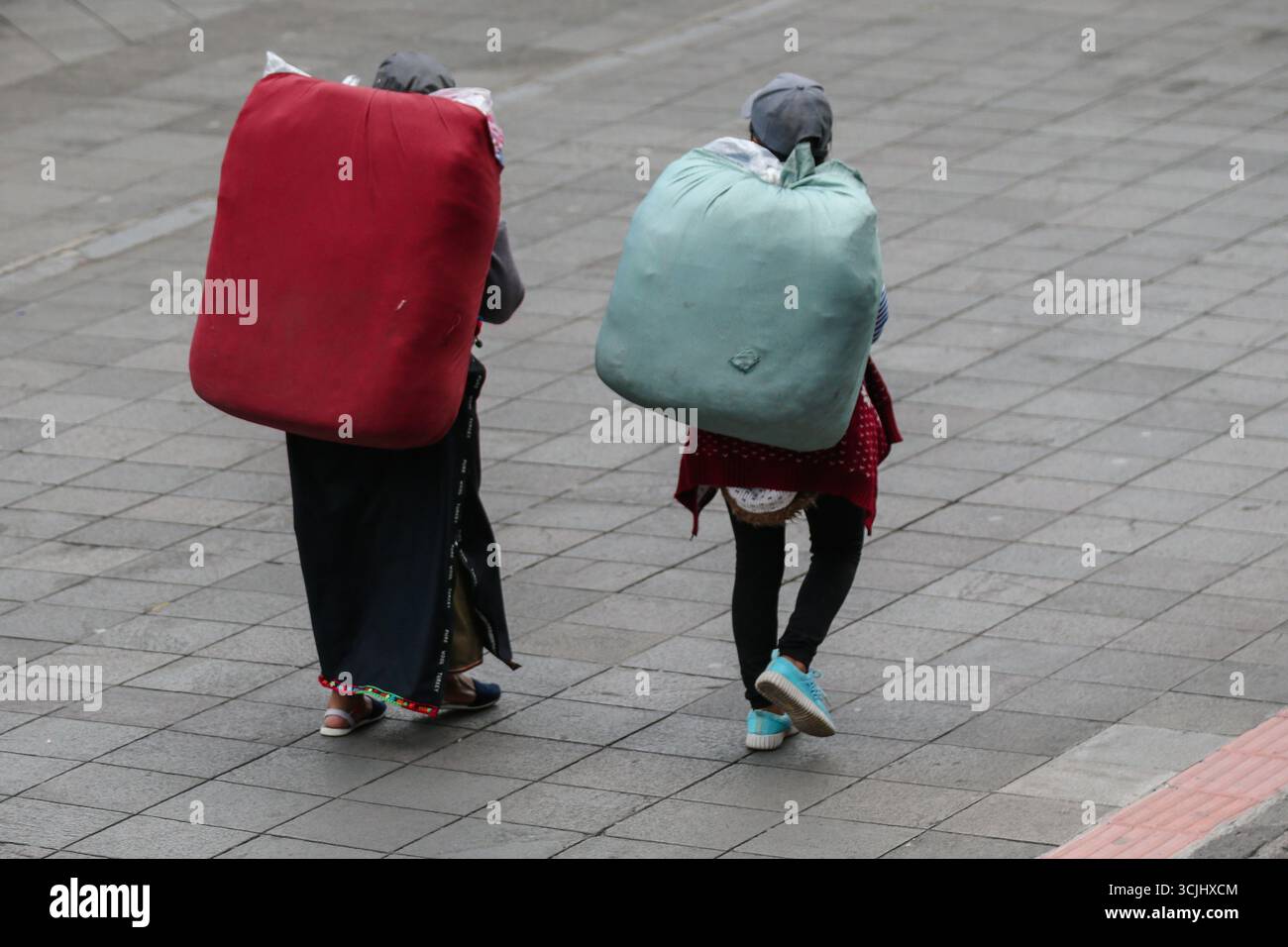 Quito, Ecuador - 23 marzo 2018: Due donne indigene camminano attraverso una strada urbana lastricata di pietra a Quito portando grandi fasci di tessuti sulle spalle Foto Stock