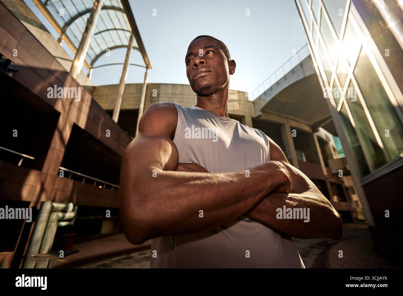 Un giovane atleta determinato e sicuro di sé si trova all'aperto con le braccia incrociate. Lo sfondo presenta un'architettura urbana accentuata dalla calda luce del sole, c Foto Stock