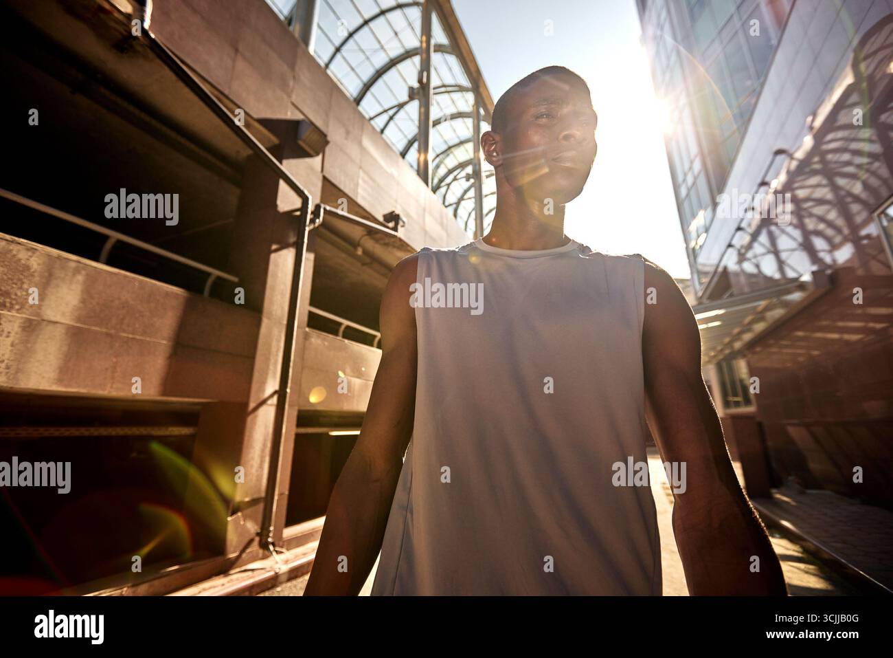 Un uomo che indossa una camicia senza maniche cammina in un ambiente urbano durante una giornata di sole. La scena presenta elementi architettonici moderni, con luce solare Foto Stock