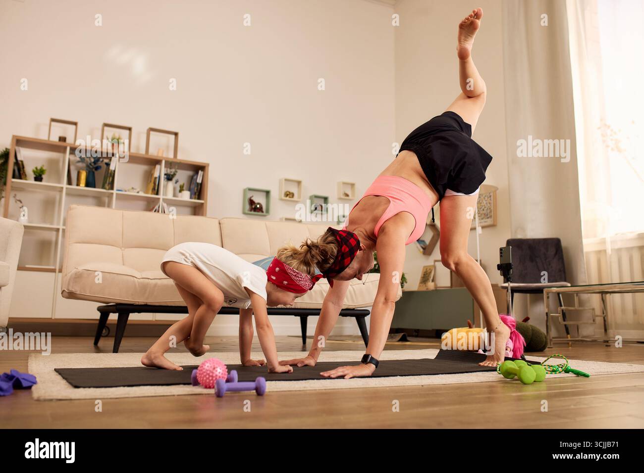 Un genitore e suo figlio si impegnano in esercizi di yoga nel loro salotto accogliente e ben illuminato. I due praticano le loro pose sui tappetini da ginnastica circondati da vibr Foto Stock