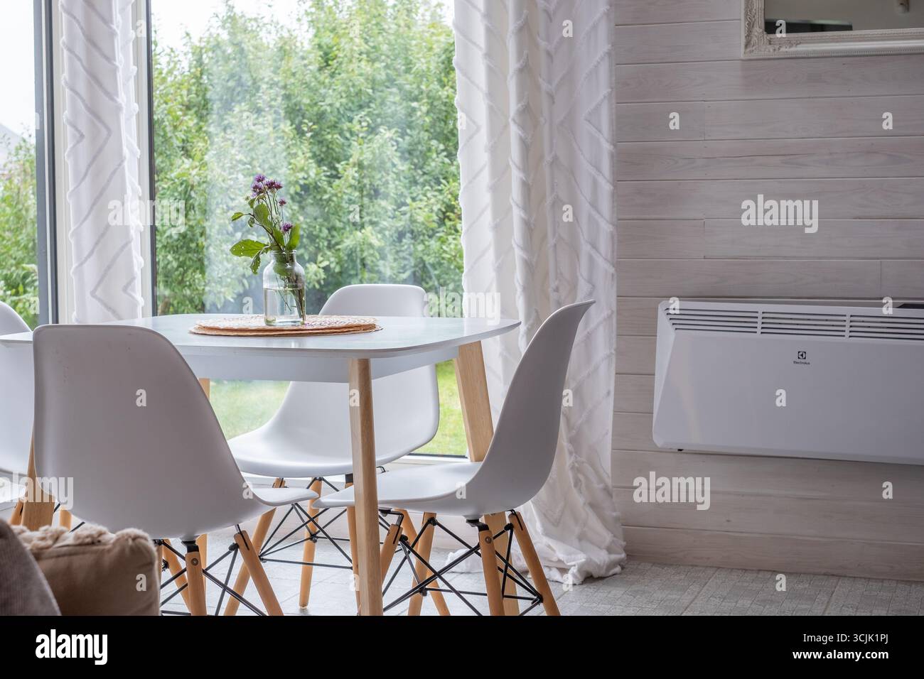 Zona pranzo in stile scandinavo con un tavolo rotondo e sedie bianche, circondata da grandi finestre, a creare un ambiente luminoso e arioso pieno di natur Foto Stock