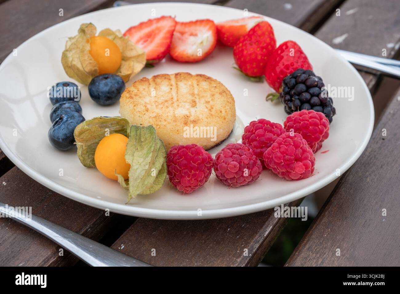 Vassoio di pancake al formaggio colorato con fragole, mirtilli, lamponi e una torta dorata su un piatto bianco, enfatizzando il cibo fresco e sano Foto Stock