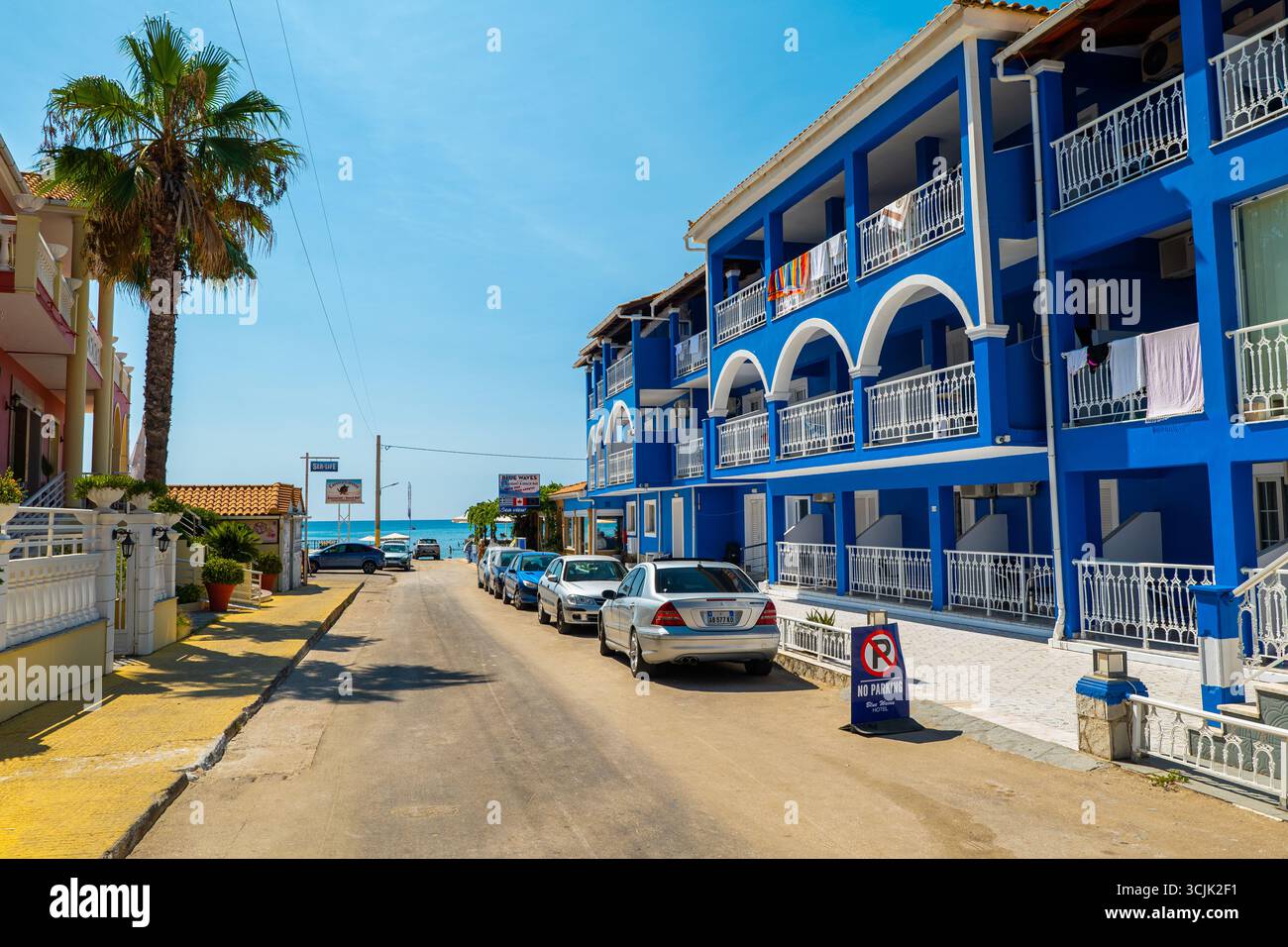 Strada a Laganas, isola di Zante, Grecia, con hotel blu che costruisce palme e auto parcheggiate che conducono verso il mare, popolare luogo di viaggio estivo Foto Stock Strada a Laganas, isola di Zante, Grecia, con hotel blu che costruisce palme e auto parcheggiate che conducono verso il mare, popolare luogo di viaggio estivo Foto Stock