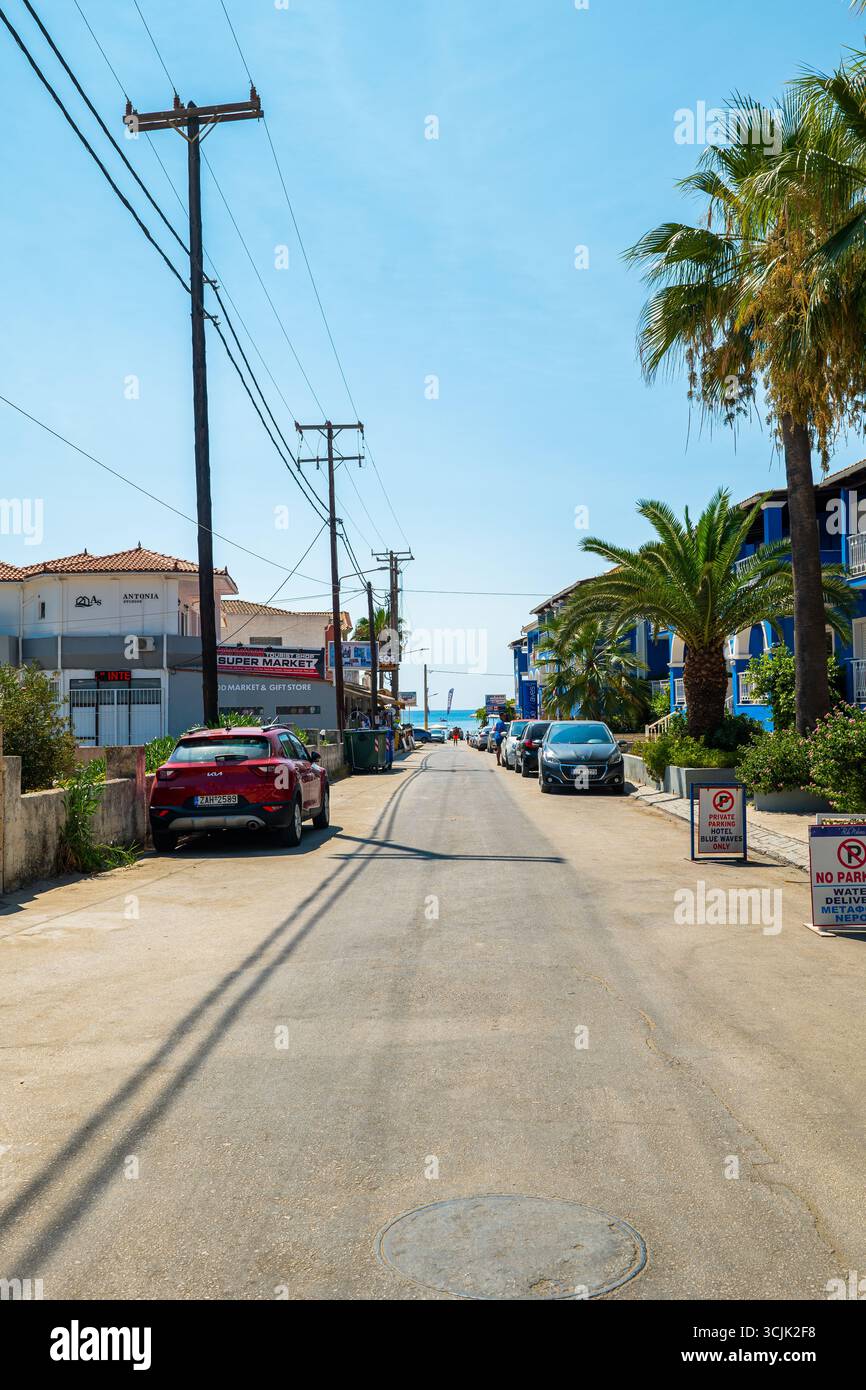 Laganas, isola di Zante, vista sulla strada della Grecia, con edifici colorati, auto lungo la strada e mare visibili alla fine della movimentata località estiva Foto Stock Laganas, isola di Zante, vista sulla strada della Grecia, con edifici colorati, auto lungo la strada e mare visibili alla fine della movimentata località estiva Foto Stock