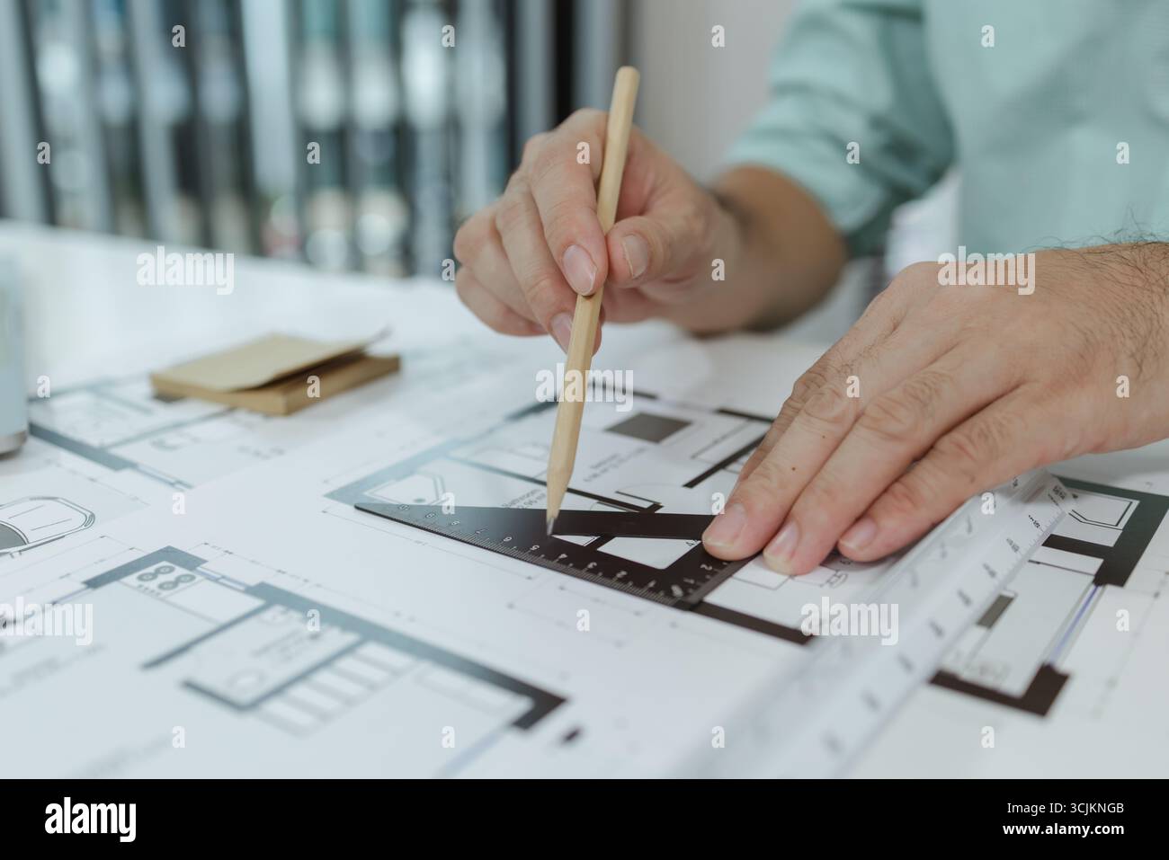 Un architetto utilizza un righello a triangolo in metallo per disegnare linee precise sui progetti della casa disposti su una scrivania bianca, concentrandosi sulla precisione e sui dettagli durante il wor Foto Stock