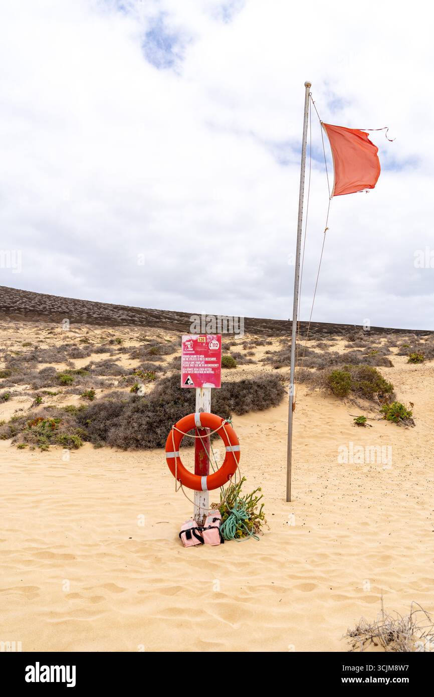Una bandiera rossa su una spiaggia indica condizioni pericolose, accanto alle attrezzature salvavita in un ambiente costiero sabbioso. Foto Stock