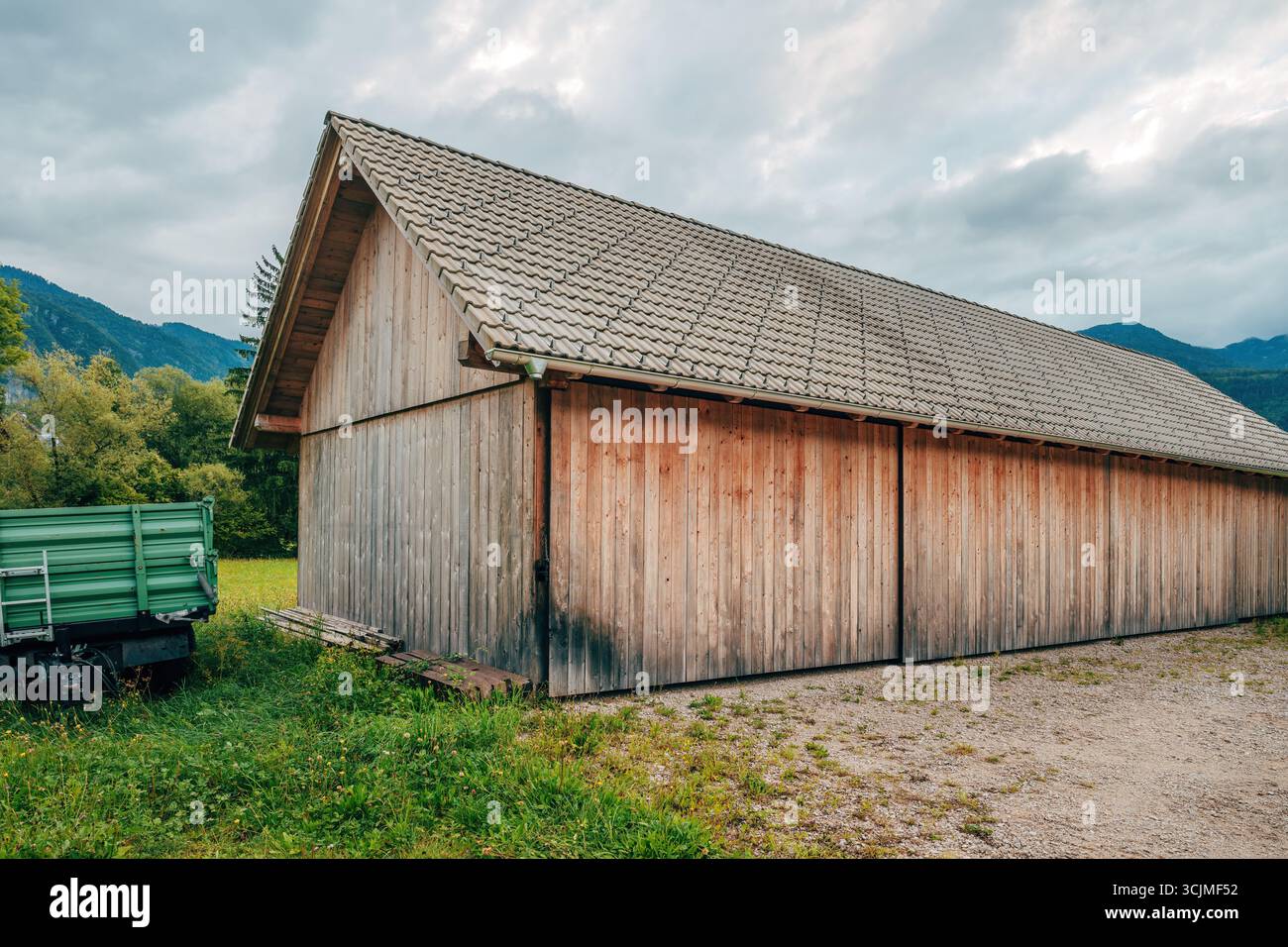 Fienile in legno con tetto piastrellato e roulotte verde nella Slovenia rurale, circondato da campi e montagne. Messa a fuoco selettiva. Foto Stock