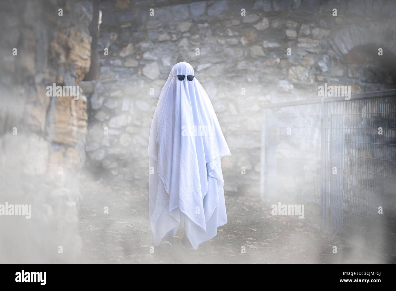 Un bambino vestito da fantasma con gli occhiali da sole cammina attraverso il fumo bianco. Halloween Masquerade Foto Stock