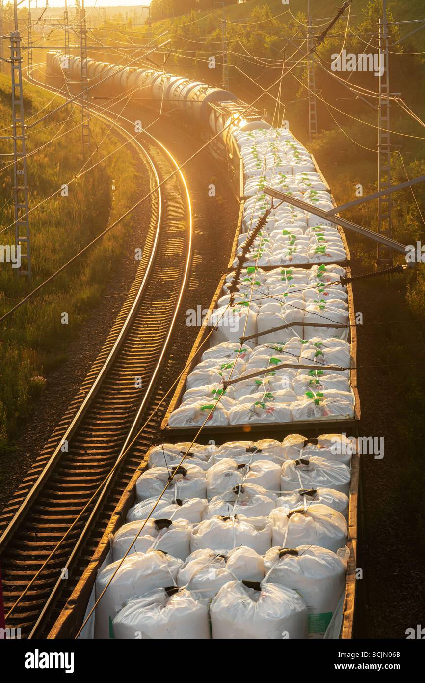 Un treno con vagoni carichi di sostanze chimiche o fertilizzanti minerali in balle e borse viaggia al tramonto lungo i binari ferroviari. Mandata fertilizzante e. Foto Stock