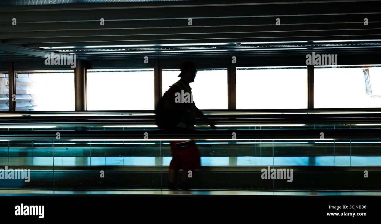 Silhouette di uomo irriconoscibile su un passaggio pedonale o una scala mobile all'interno della sala del terminal dell'aeroporto. Visione grandangolare, retroilluminazione, luce soffusa Foto Stock