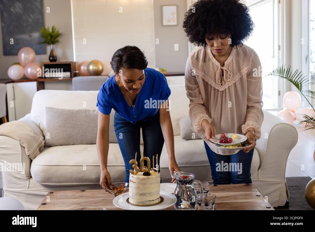 Donne che preparano i festeggiamenti di compleanno con torta e cesto di frutta nel soggiorno Foto Stock