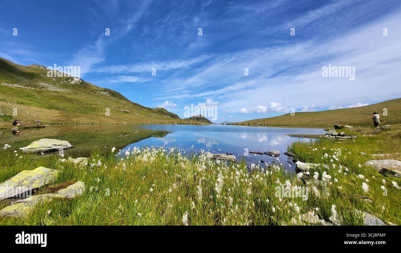 Un idilliaco lago di montagna nelle Alpi svizzere con erba di cotone lungo la riva e nuvole mostruose che scivolano attraverso un cielo blu profondo Foto Stock