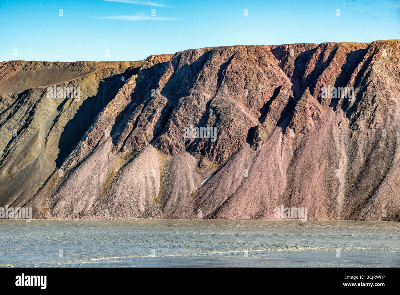 Paesaggio di Faksevagen Spitsbergen Svalbard Norvegia // SPITSBERGEN, Svalbard e Jan Mayen - il paesaggio di Faksevagen presenta montagne aspre e multicolore con pendii ripidi e pendii ricoperti di raso. Il terreno presenta diversi strati geologici, con formazioni rocciose grigie più scure che passano in tonalità rossastro-bruno e rossastro, indicativi di diverse composizioni minerali. Un corpo d'acqua, forse un fiordo o una baia, si trova alla base delle montagne, riflettendo il cielo azzurro. Questa regione artica è conosciuta per la sua spettacolare e cruda bellezza naturale, modellata dall'attività glaciale e dal permafrost. Foto Stock