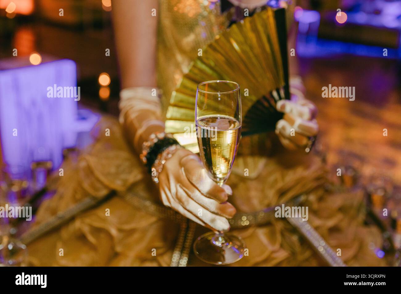 Una donna in abito dorato si gode una bevanda frizzante mentre tiene elegantemente un fan in un'atmosfera da festa vivace. Foto Stock
