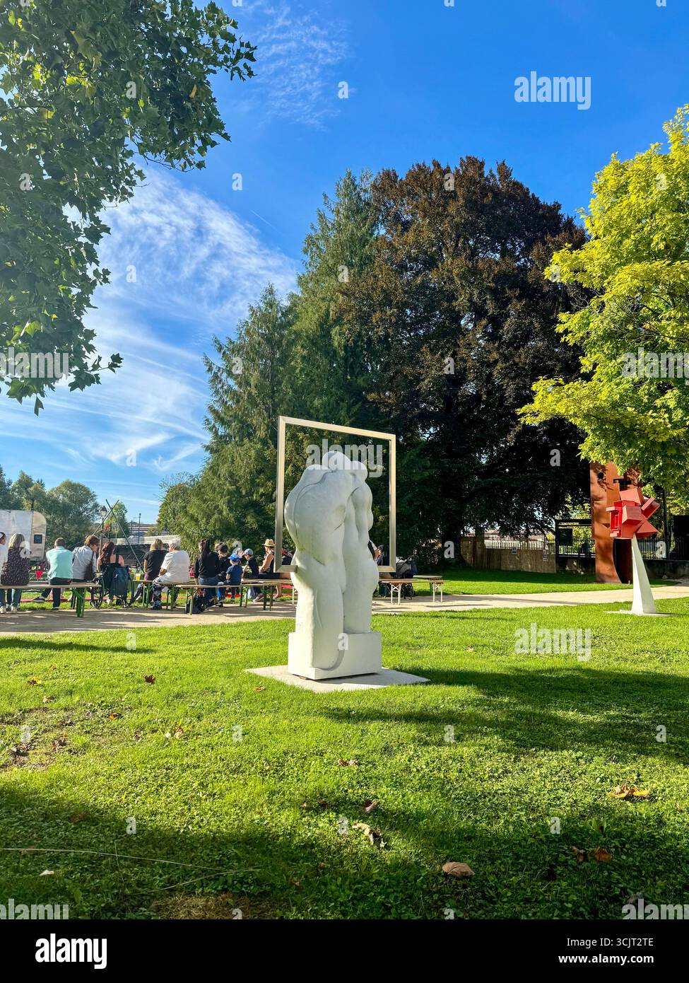 Moderna scultura bianca in un parco pubblico durante l'evento culturale all'aperto, con persone sedute sullo sfondo sotto gli alberi e il cielo blu - Immagine stock catturata con smartphone