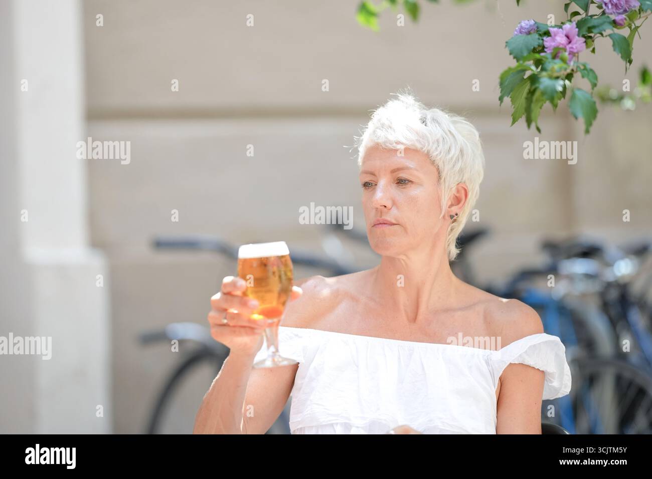 Una donna è seduta fuori con un bicchiere di birra in mano. Indossa una camicia bianca e i capelli vengono tirati indietro. La scena è rilassata e casua Foto Stock