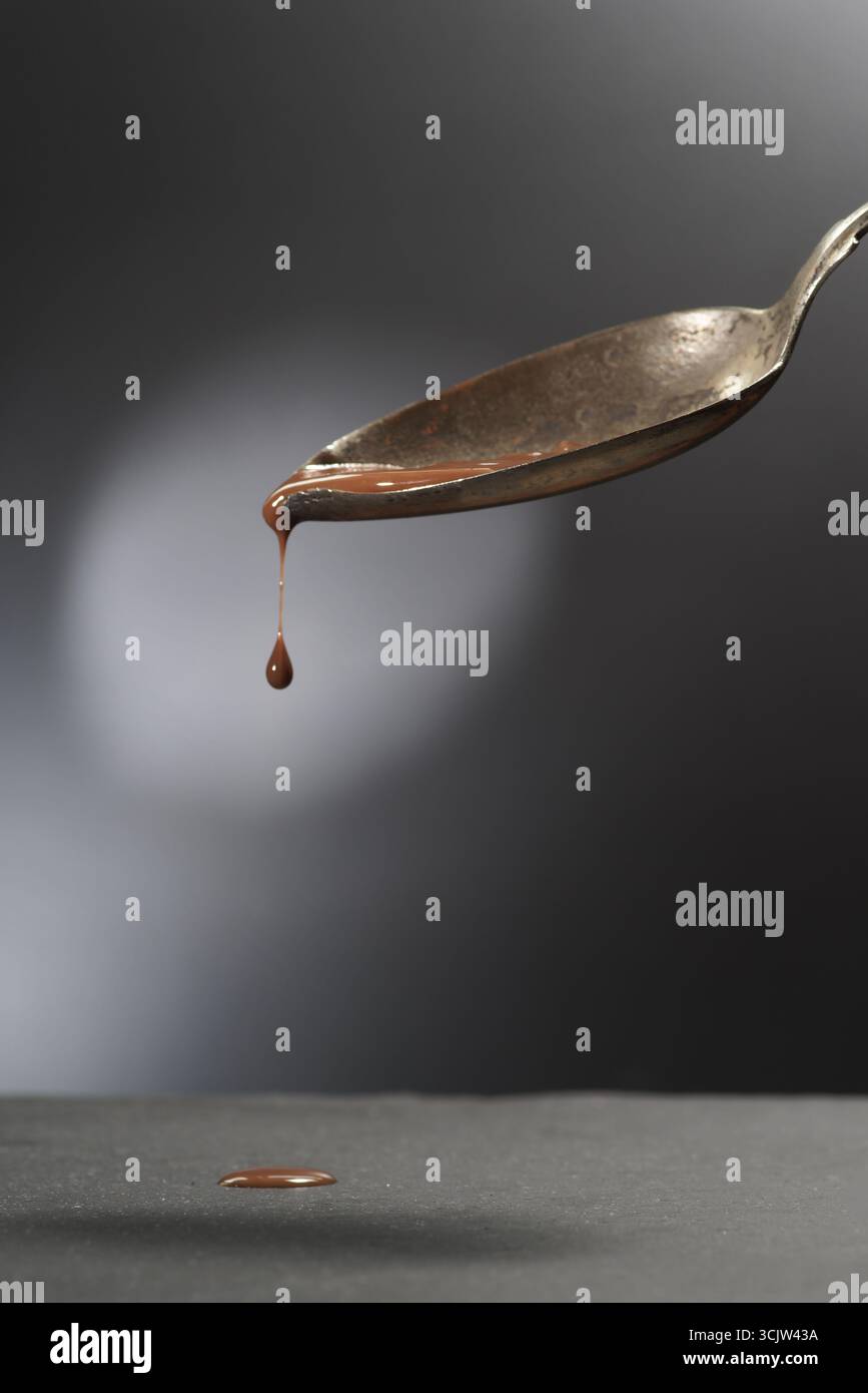 Il cioccolato liquido dolce scorre da un cucchiaio di metallo inclinato su uno sfondo scuro Foto Stock