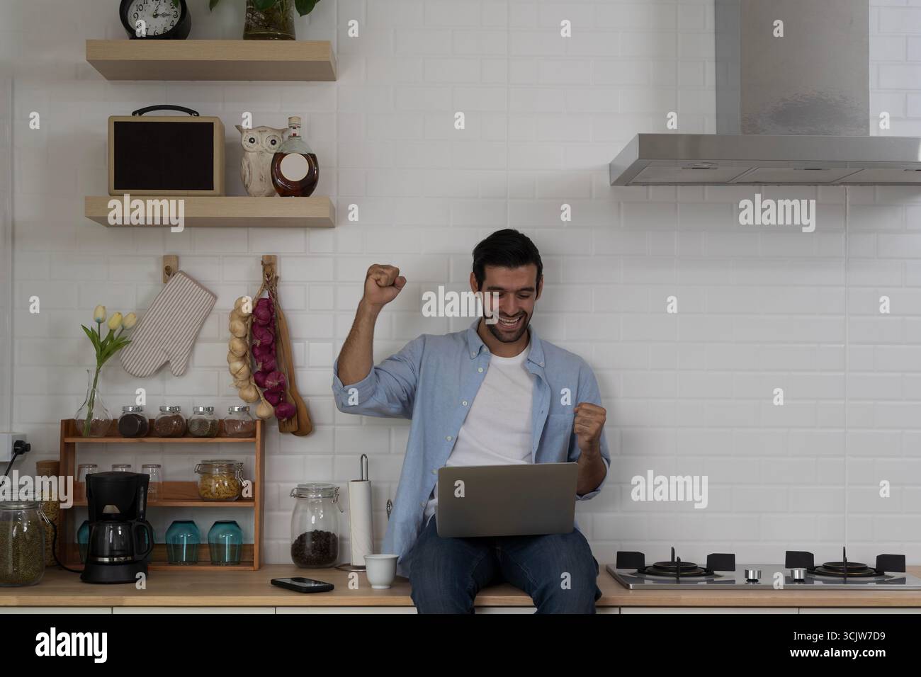 Giovane eccitato seduto sul bancone della cucina che celebra il successo con il laptop, sorride e solleva pugni in uno stato d'animo gioioso, il concetto di lavoro a distanza raggiunge Foto Stock