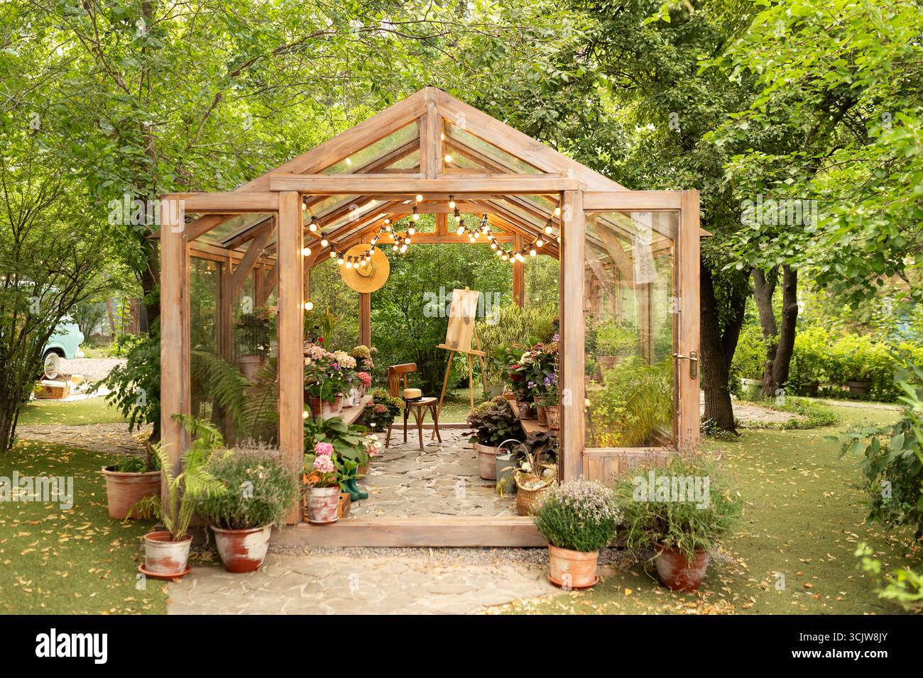 Accogliente giardino con serra in vetro e sedia. Interno di una serra illuminata in legno con mobili, piante e fiori in vasi sul retro. Foto Stock