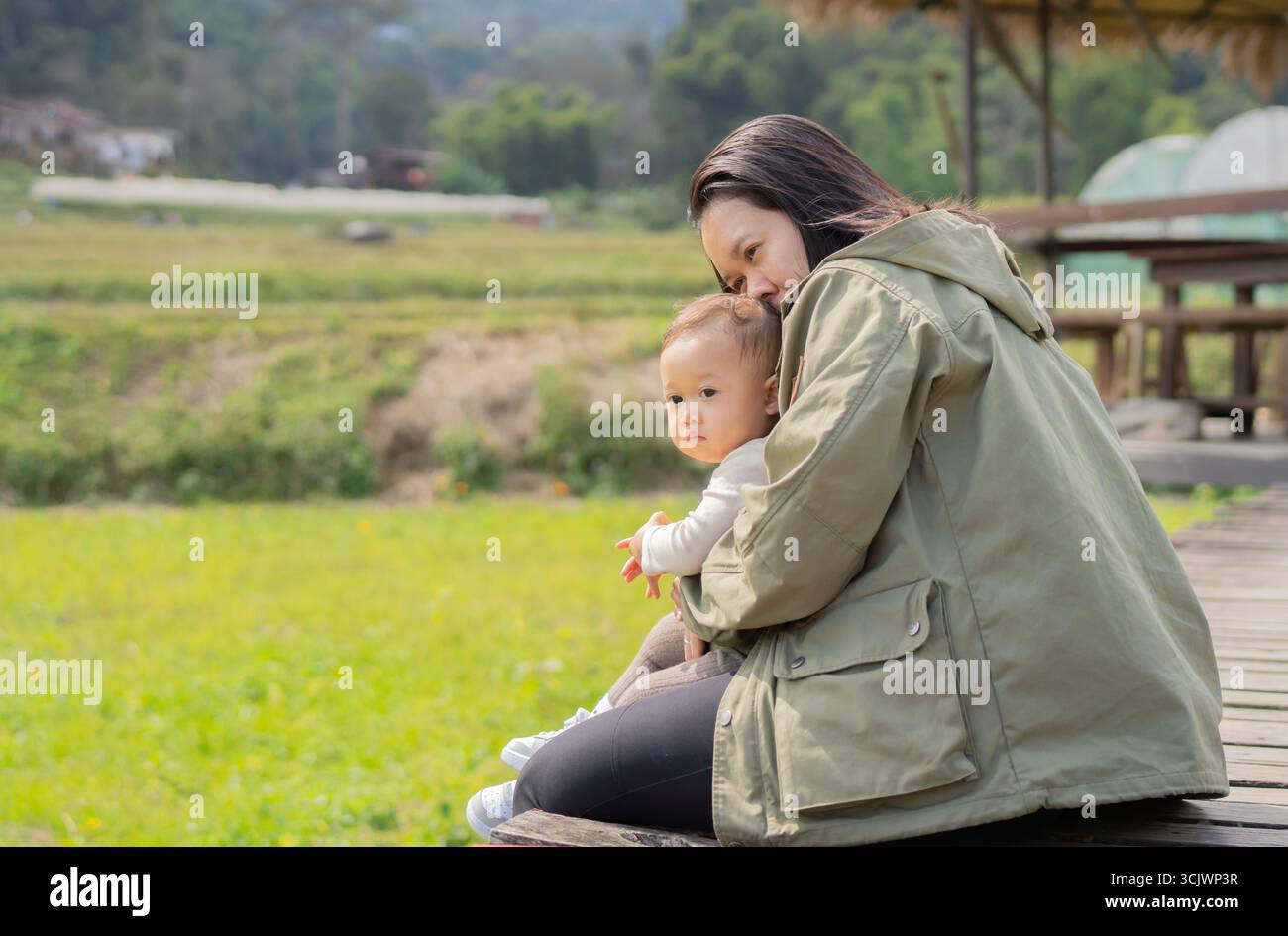 Felice madre asiatica abbraccia il suo bambino seduto nel giardino naturale al tramonto. La famiglia trascorre del tempo insieme all'aria aperta. Foto Stock