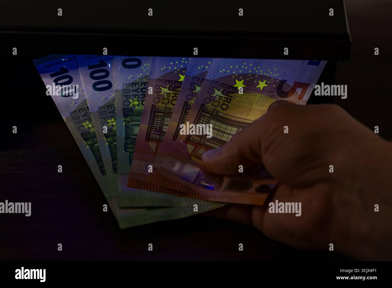 Un uomo tiene in mano le banconote in euro e le controlla per verificarne l'autenticità sotto la luce ultravioletta, da vicino. Segnali ed elementi di sicurezza sui reali 100 e. Foto Stock