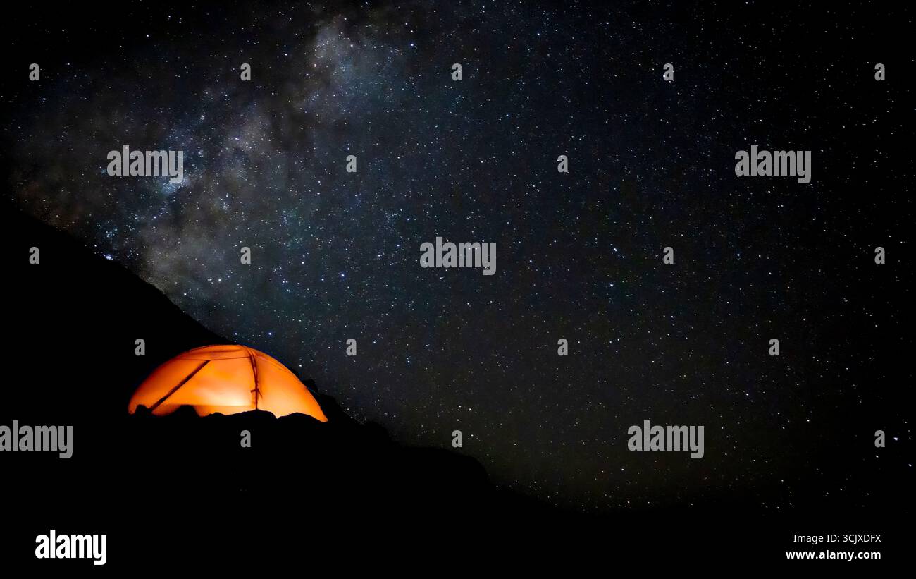 Una tenda illuminata di arancio si illumina di calore nell'oscurità alla base di una collina. È l'unico segno di presenza umana in un vasto e selvaggio paesaggio. Il cielo sopra Foto Stock