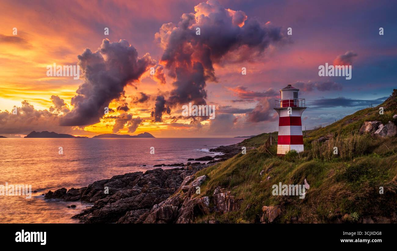 Un faro a strisce rosse e bianche sorge su una scogliera rocciosa e verde che si affaccia su un mare calmo. Il cielo sopra è uno spettacolo mozzafiato di un tramonto, con Foto Stock