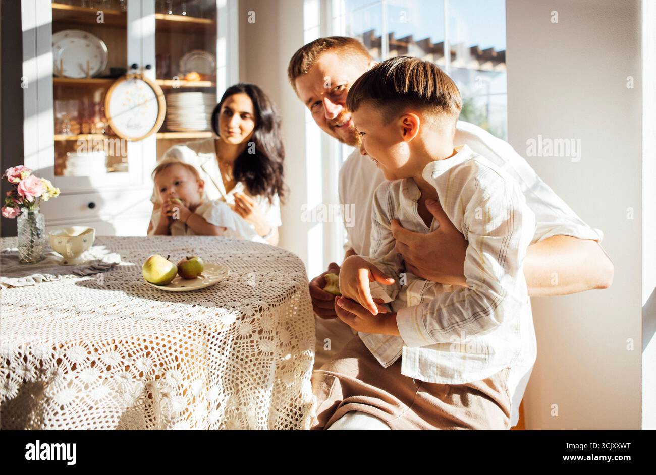 La famiglia si è riunita in una cucina illuminata dal sole, condividendo momenti di gioia intorno a un tavolo, con texture morbide e un ambiente accogliente che esalta la loro connessione e la loro happ Foto Stock