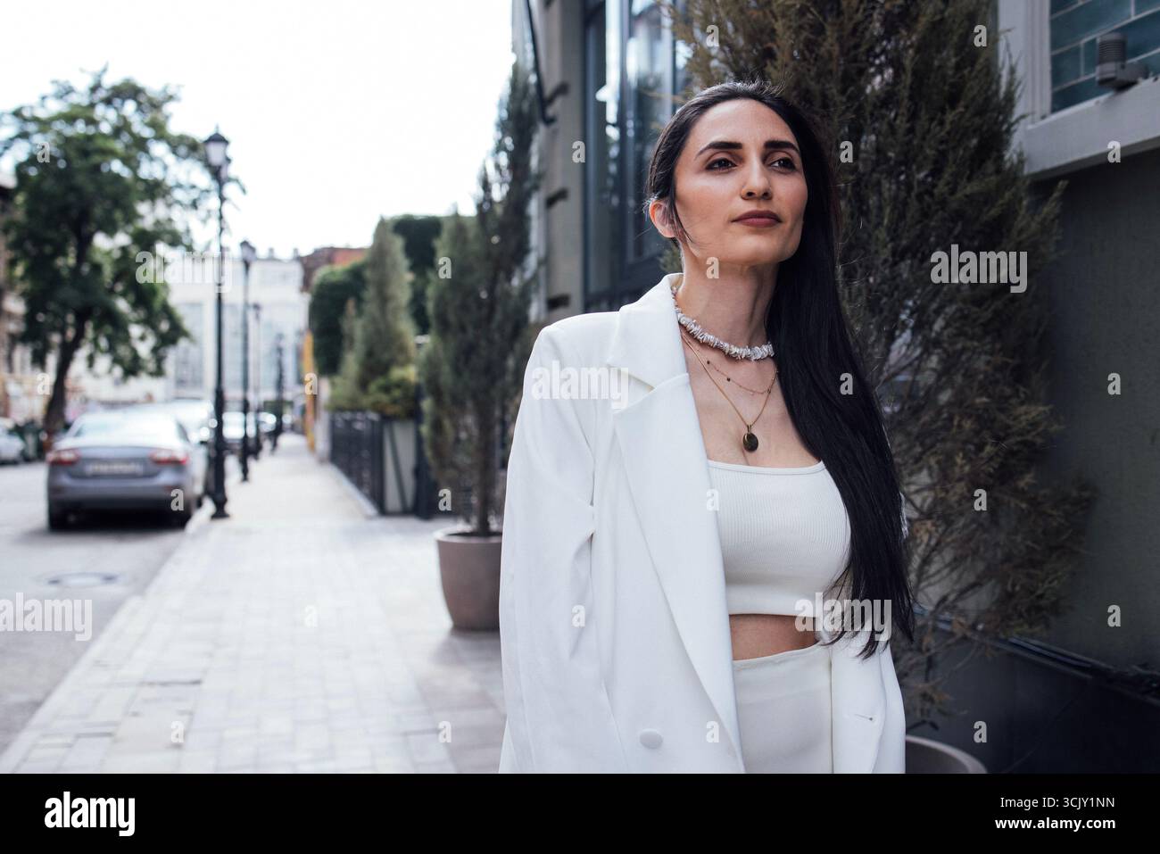 Una donna elegante vestita di bianco si erge in modo sicuro sulla strada della città, circondata dal verde e dall'architettura urbana, incarnando la moda contemporanea Foto Stock