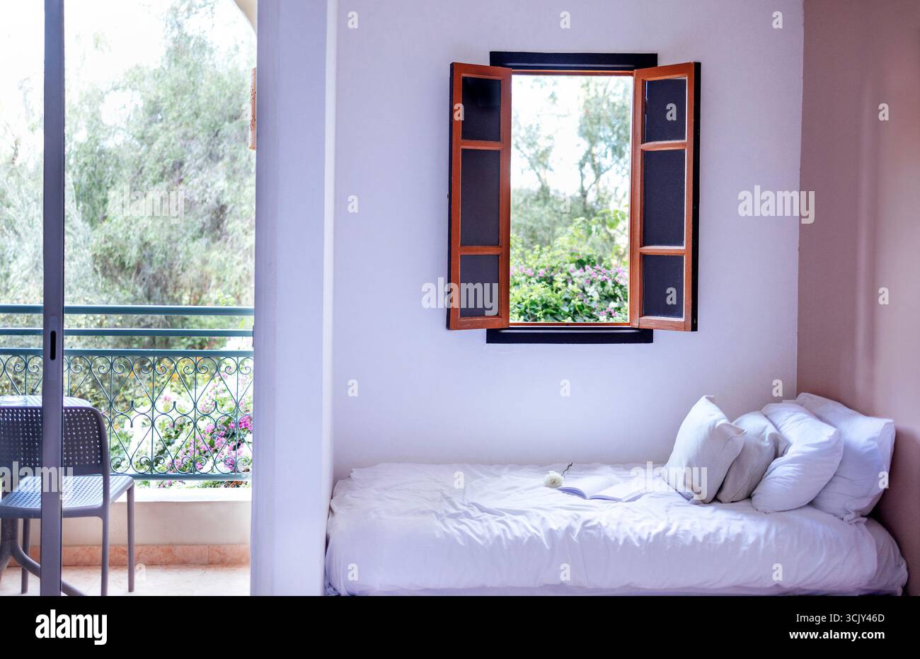 L'invitante camera da letto presenta una finestra aperta con vegetazione esterna, un letto ben fatto con lenzuola e cuscini bianchi, creando un ambiente tranquillo e accogliente Foto Stock