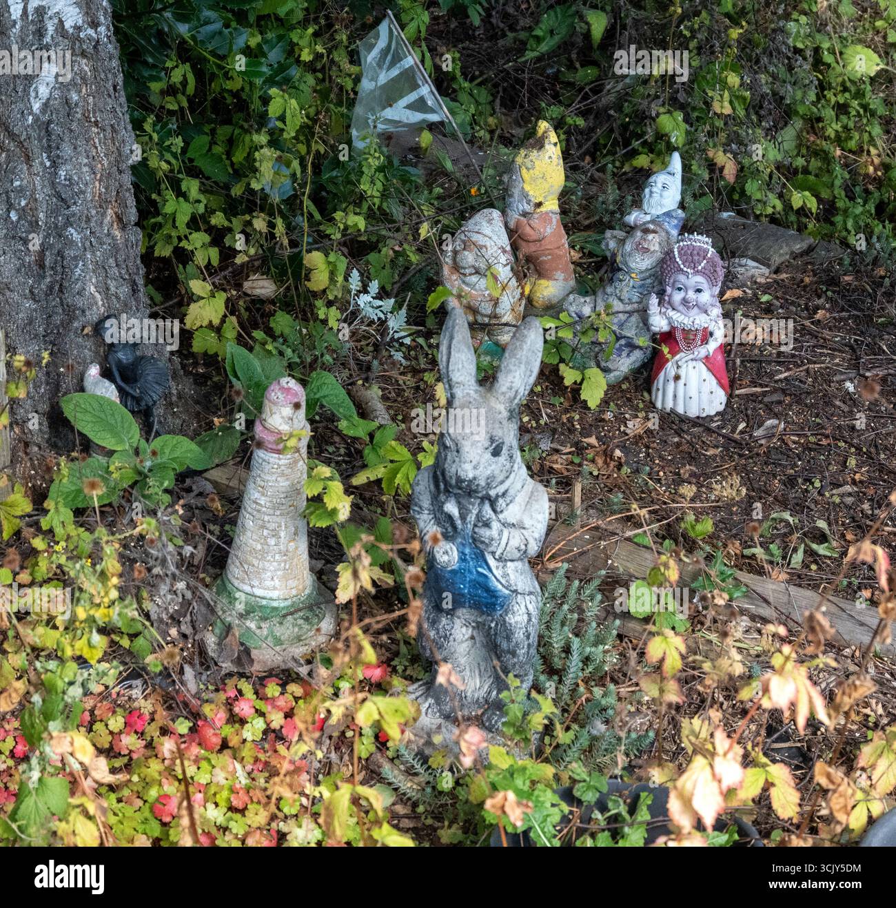 Una collezione di ornamenti da giardino vintage, tra cui gnomi e una statuetta di coniglio, annidata tra piante e fogliame in un angolo ombreggiato del giardino Foto Stock