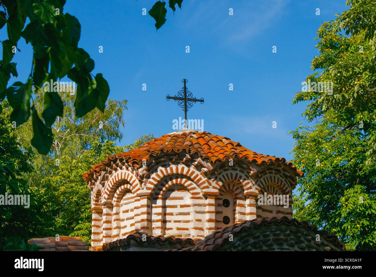 Tradizionale cupola ortodossa con tegole rosse in terracotta e croce metallica decorativa, incorniciata da alberi verdi e cielo azzurro limpido in un'estate soleggiata Foto Stock