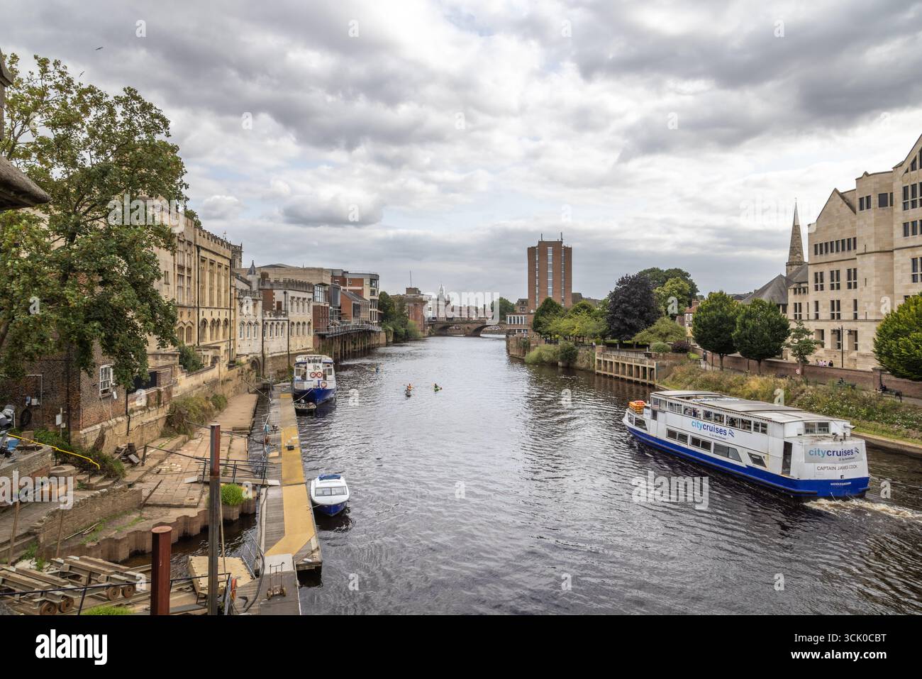 Barche e kayak navigano sul fiume Ouse a York, Inghilterra, fiancheggiato da edifici storici e moderni, catturando il patrimonio urbano e l'attività fluviale Foto Stock