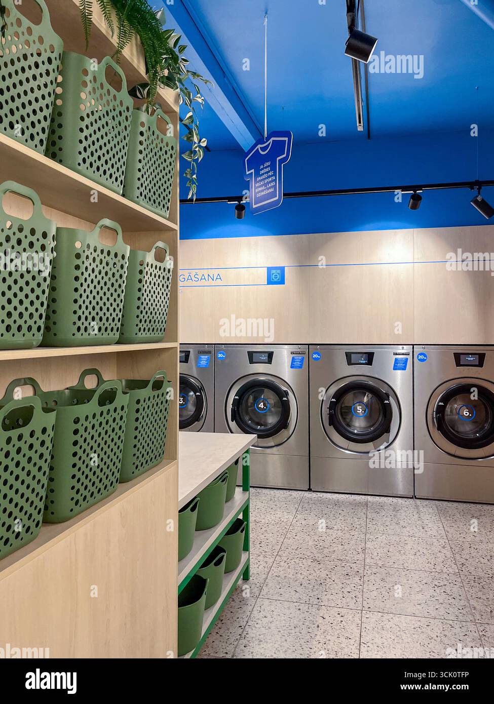 Riga, Lettonia - 4 settembre 2025: Moderna sala lavanderia con macchine Speed Queen, cesti verdi e soffitto blu luminoso che offre un servizio self-service Foto Stock
