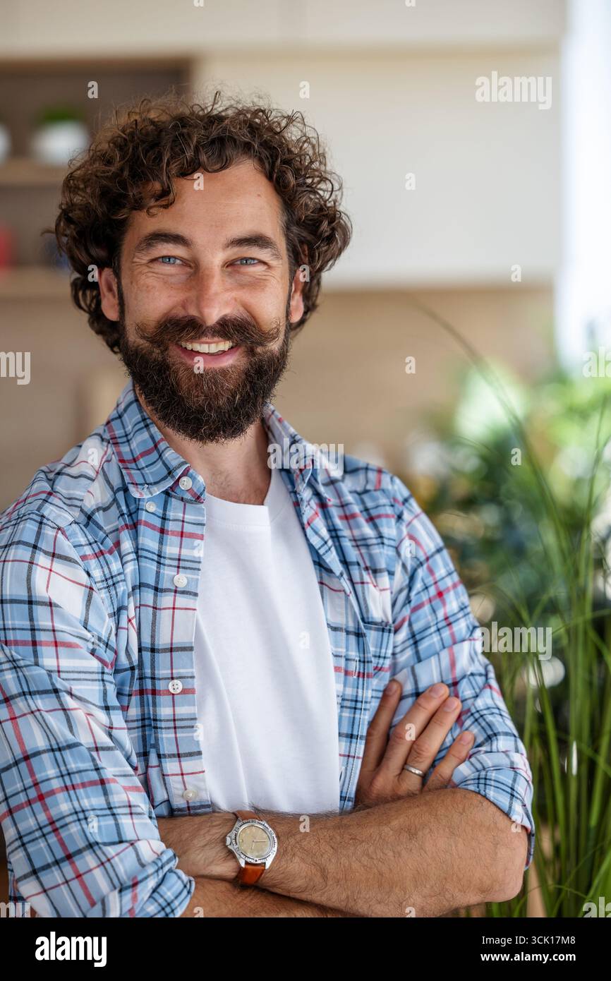 Ritratto della vista frontale di un uomo sorridente dai capelli ricci con baffi in piedi nel suo appartamento con le braccia incrociate Foto Stock