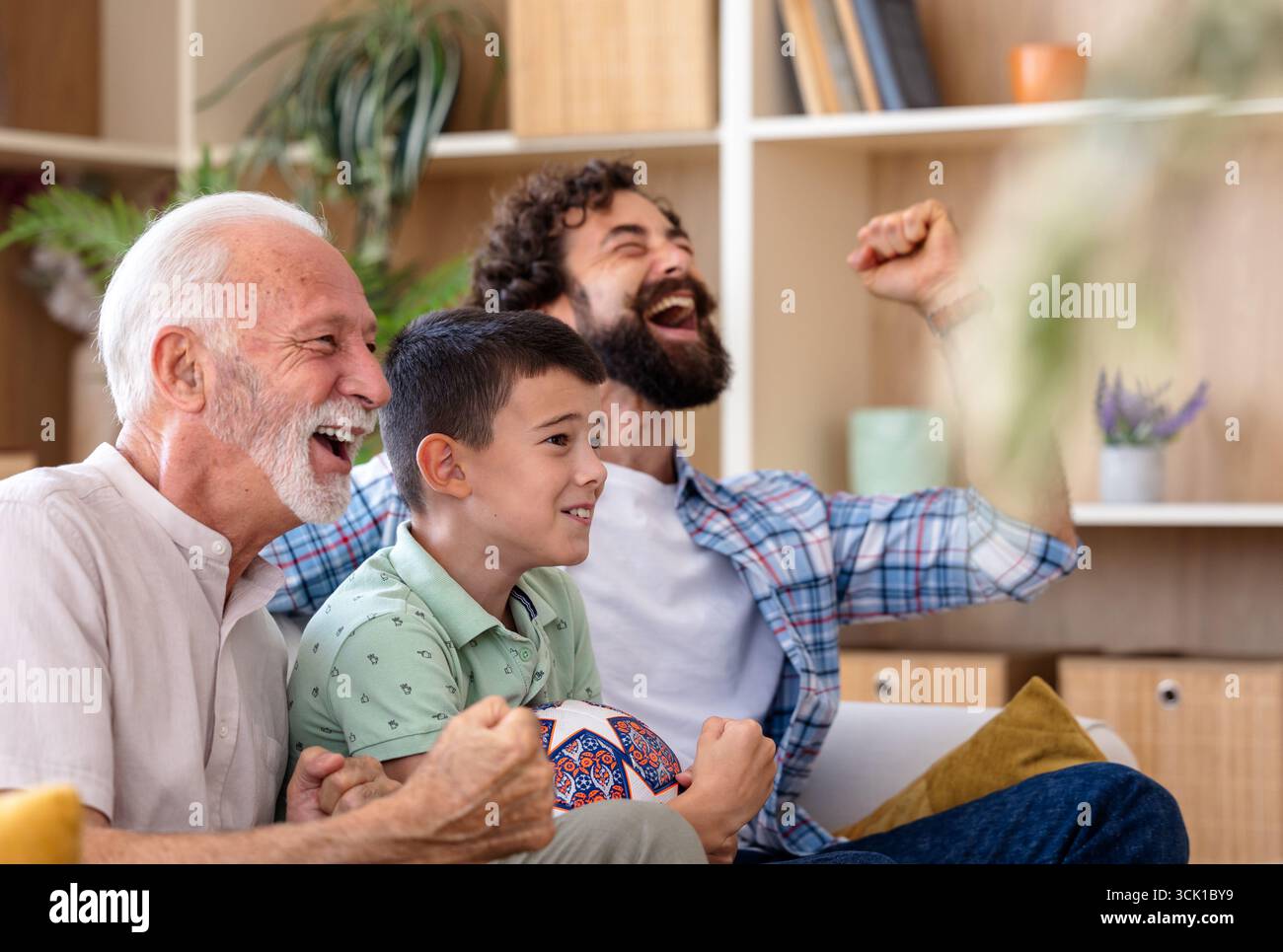 Tre generazioni condividono risate ed emozioni mentre guardano eventi sportivi in televisione in un accogliente salotto Foto Stock