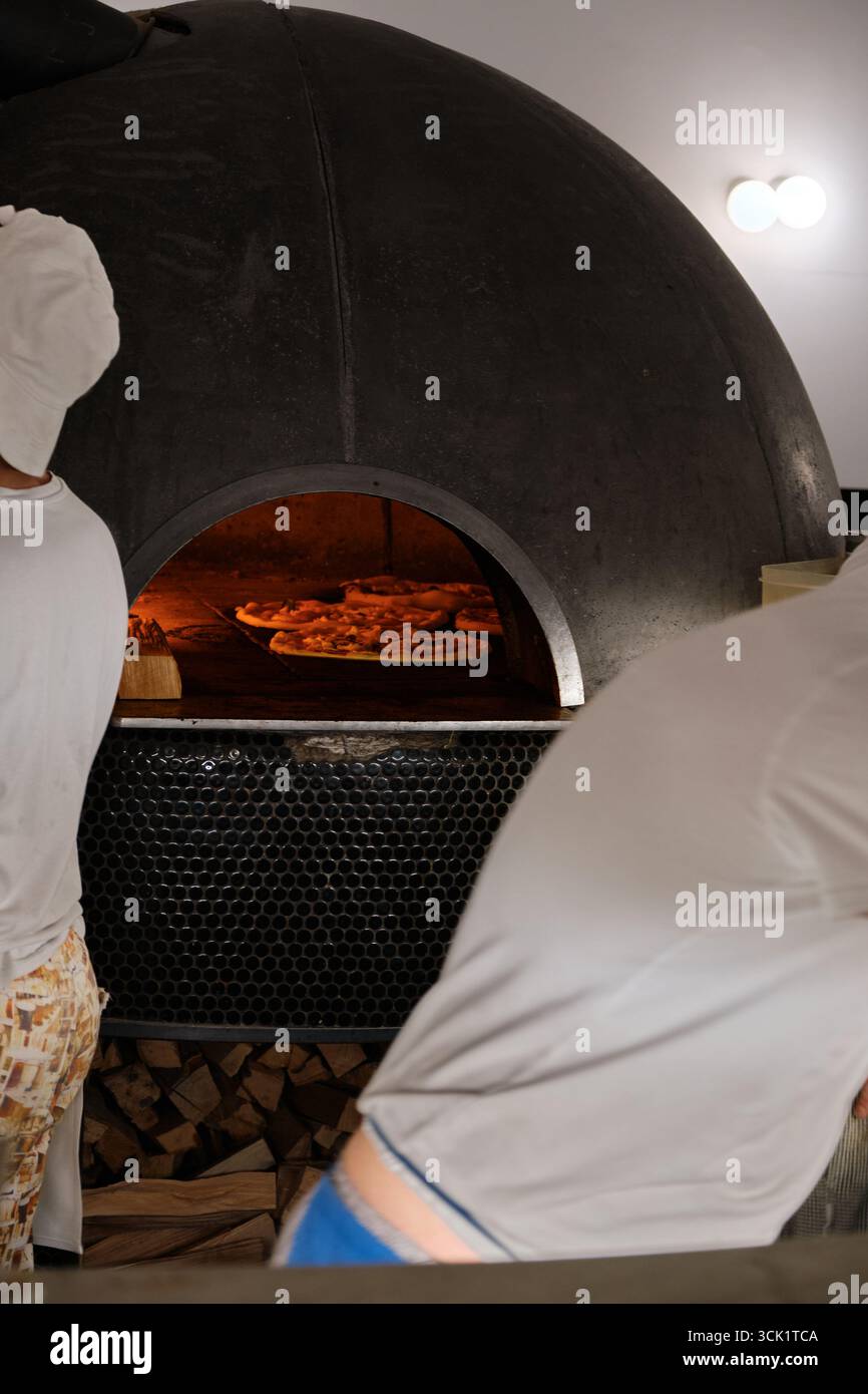 Forno per pizza semicircolare nero a legna con quattro pizze cotte all'interno e legna da ardere impilata sul fondo, che mette in mostra l'autentico ambiente della pizzeria e. Foto Stock Forno per pizza semicircolare nero a legna con quattro pizze cotte all'interno e legna da ardere impilata sul fondo, che mette in mostra l'autentico ambiente della pizzeria e. Foto Stock