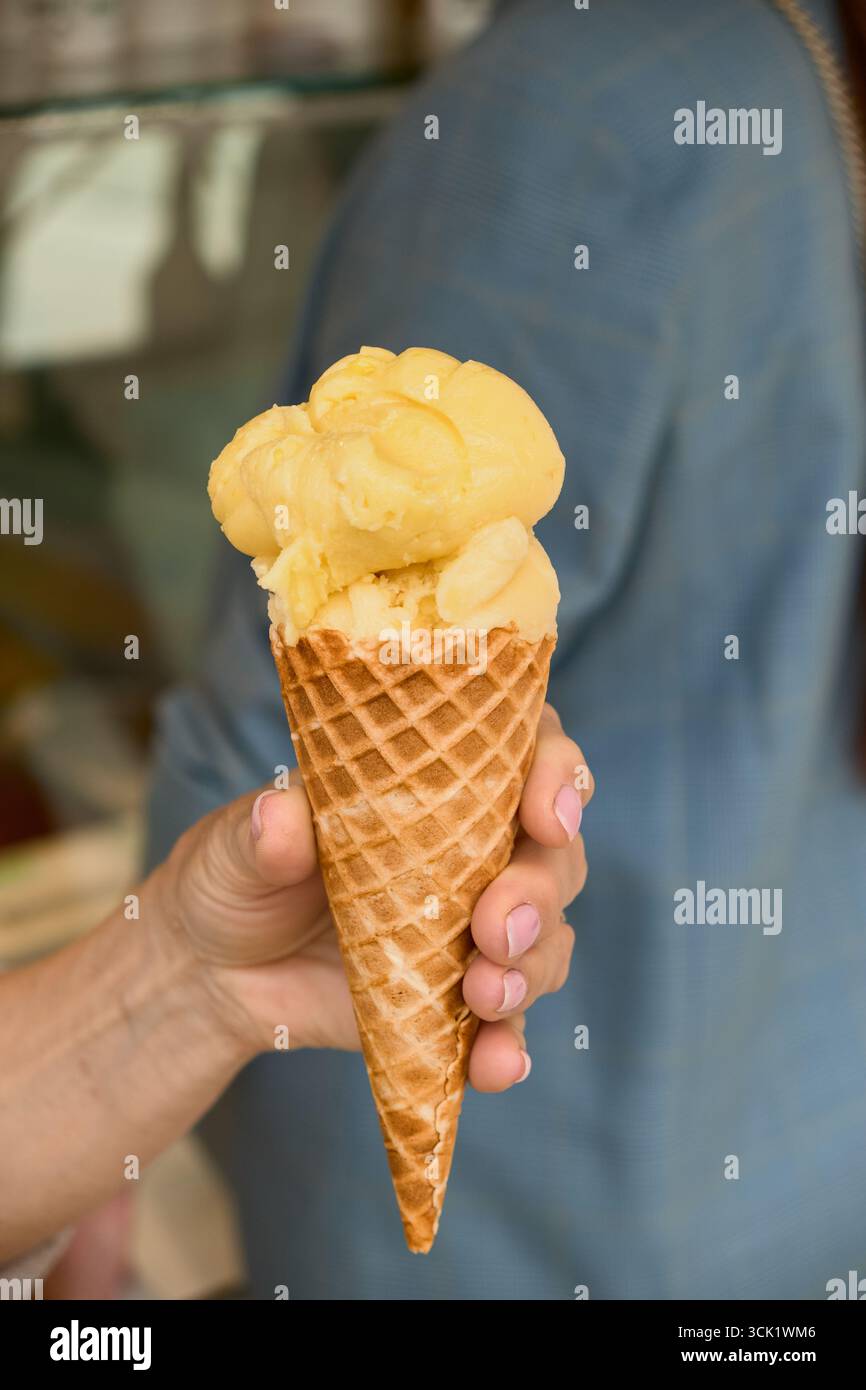 Primo piano di una mano che regge un cono di gelato al mango sullo sfondo di un blazer grigio, che mette in risalto la vibrante delizia estiva e lo stile urbano informale. Foto Stock
