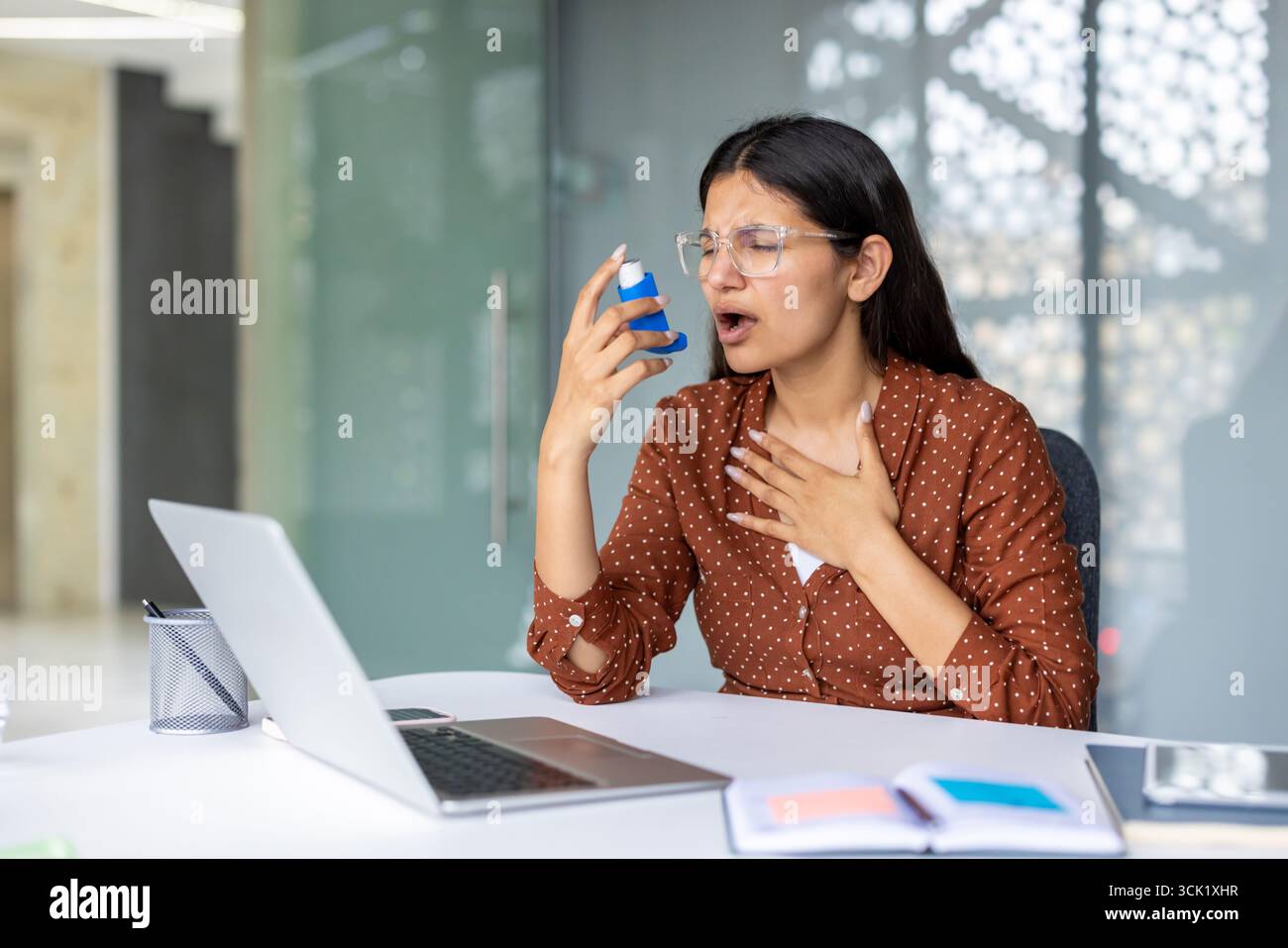 Giovane donna d'affari che soffre di asma mentre è seduta alla scrivania, utilizza un inalatore e lotta per respirare davanti al suo portatile in ufficio Foto Stock