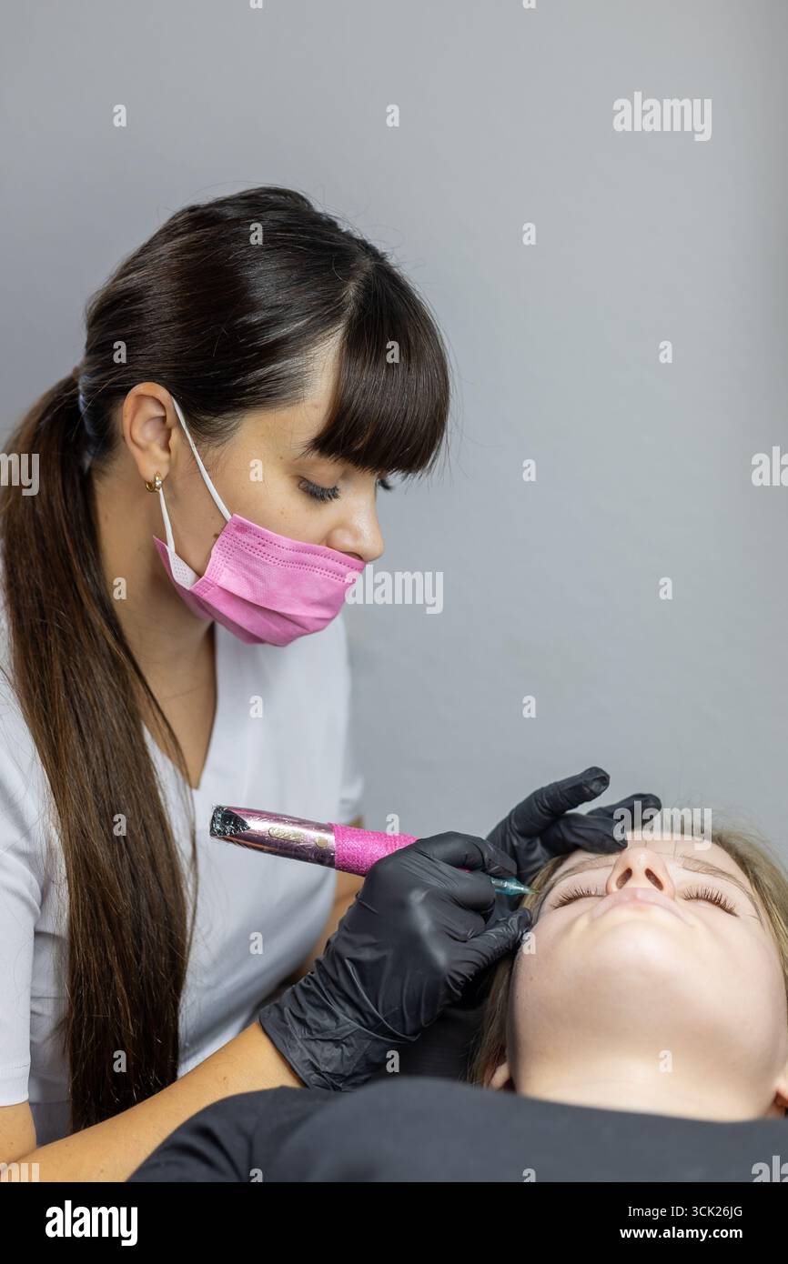 Un professionista della bellezza applica meticolosamente le tecniche di microblading alle sopracciglia del cliente in un ambiente tranquillo in studio. Il cliente si trova di nuovo conforto Foto Stock