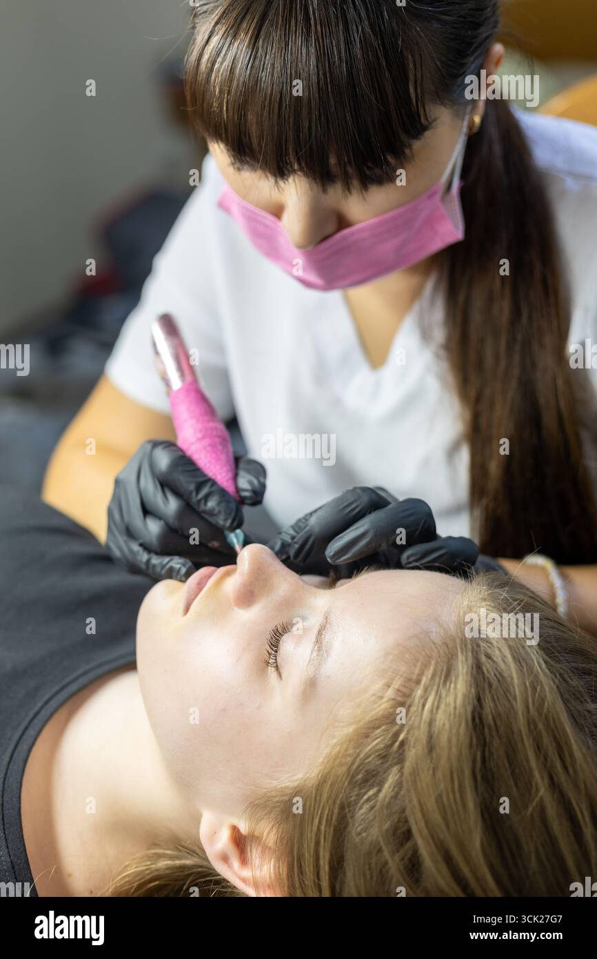 Un artista esperto sta applicando il trucco permanente delle labbra sulle labbra di un cliente in uno studio di bellezza. L'atmosfera è concentrata e professionale, garantendo precisio Foto Stock