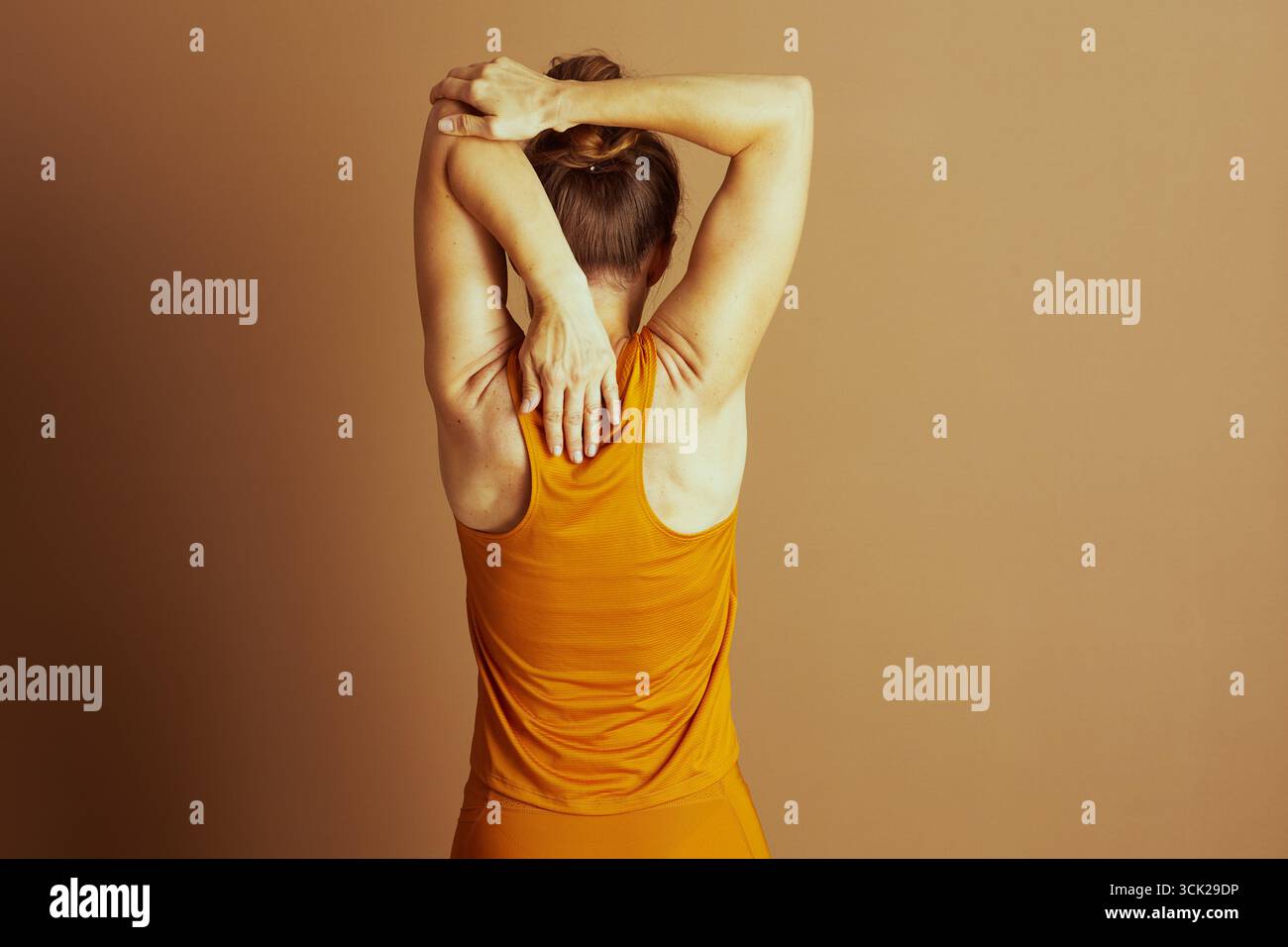 Vista dalla schiena, una donna in abbigliamento sportivo arancione si allunga la parte superiore della schiena e del braccio, le mani sulla schiena. Irradia calma durante il suo allenamento Foto Stock