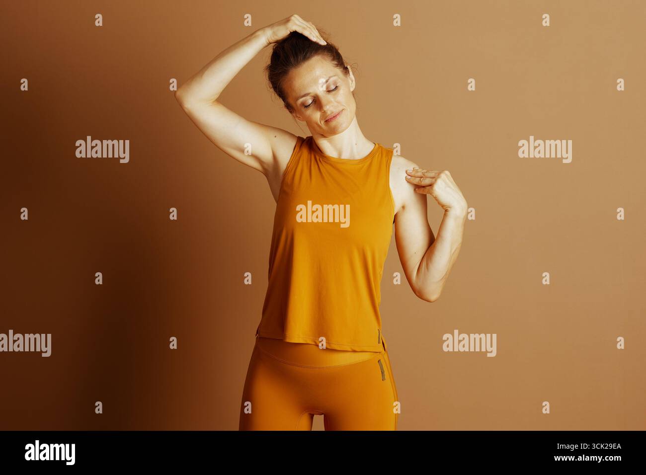 Una donna con un top arancione e leggings esegue un delicato stretching del collo, occhi chiusi, con un'espressione calma e mirata. La scena trasmette consapevolezza e. Foto Stock