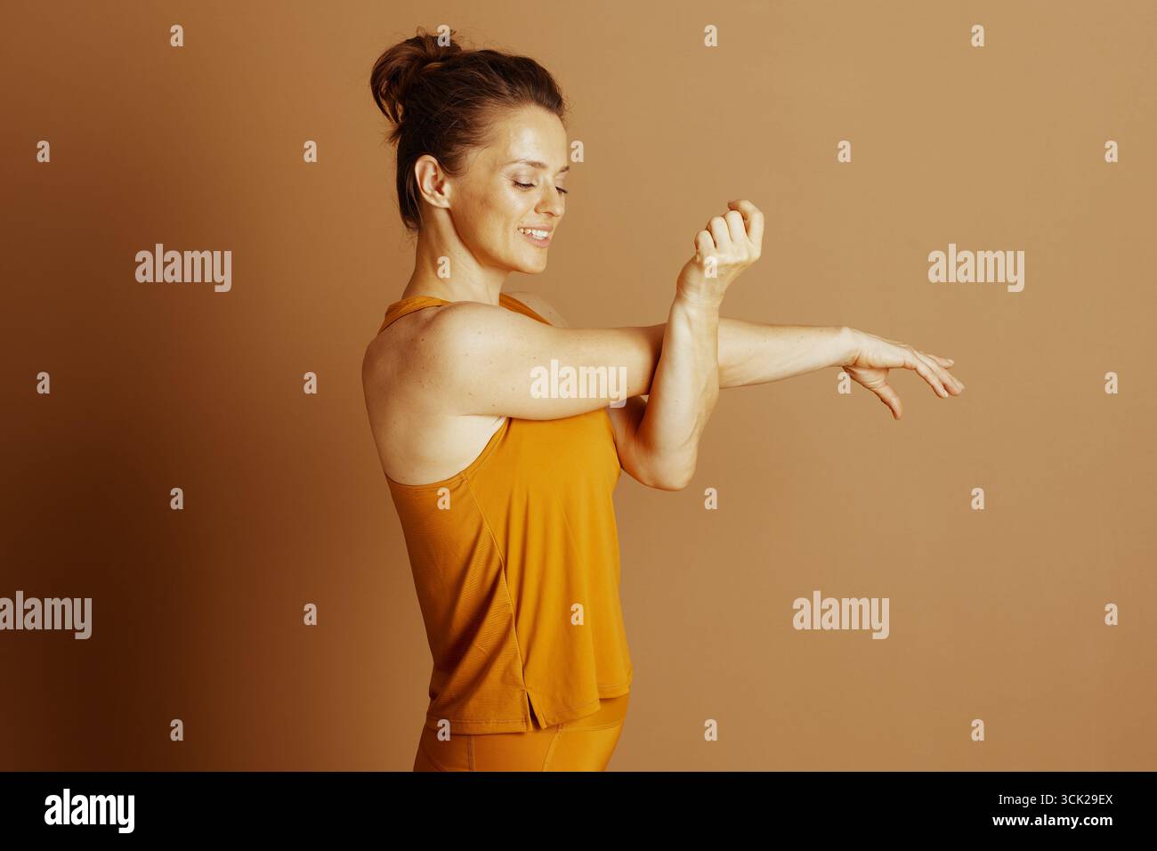 Una donna serena in abbigliamento sportivo giallo senape esegue un delicato allungamento del braccio, gli occhi chiusi a fuoco calmo. Il suo profilo laterale su un morbido sfondo marrone Foto Stock