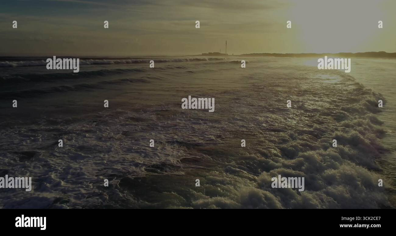 Onde ondulate e spumeggianti dell'oceano che catturano la luce dorata del tramonto sul punto panoramico della spiaggia e sul mare Foto Stock