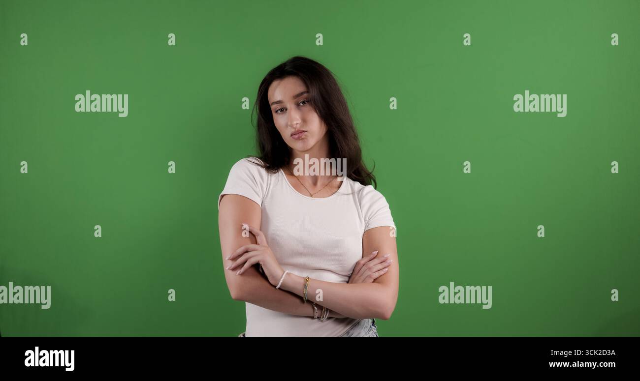 Giovane donna che posa con le braccia incrociate sullo sfondo dello schermo verde, esprimendo disaccordo e disapprovazione Foto Stock