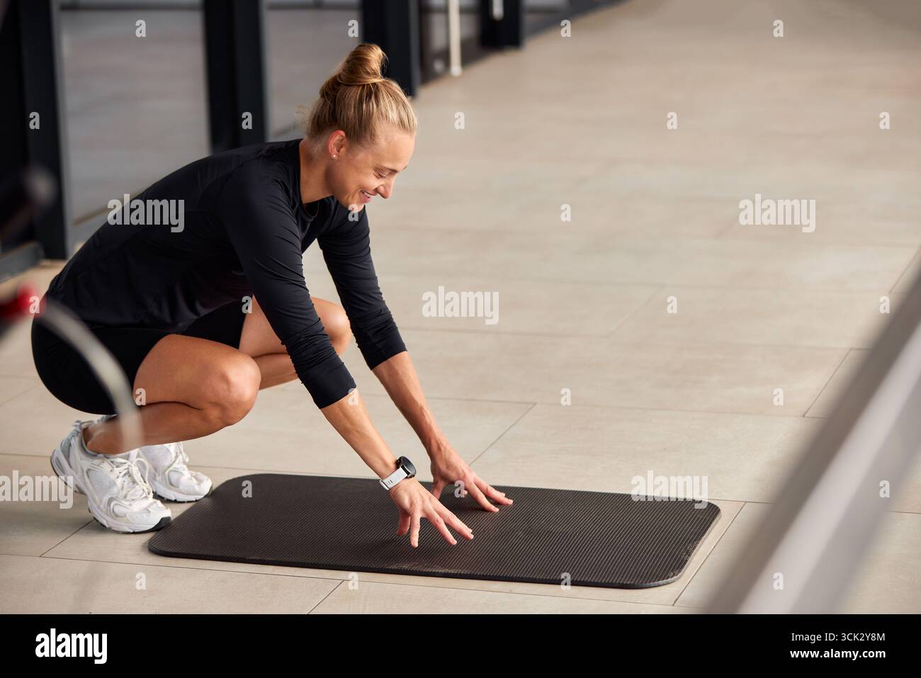 Una donna che indossa abbigliamento sportivo prepara il suo tappetino per un allenamento in un luminoso spazio interno. L'ambiente fitness e la sua posa dinamica mettono in risalto la sua attenzione alla salute Foto Stock